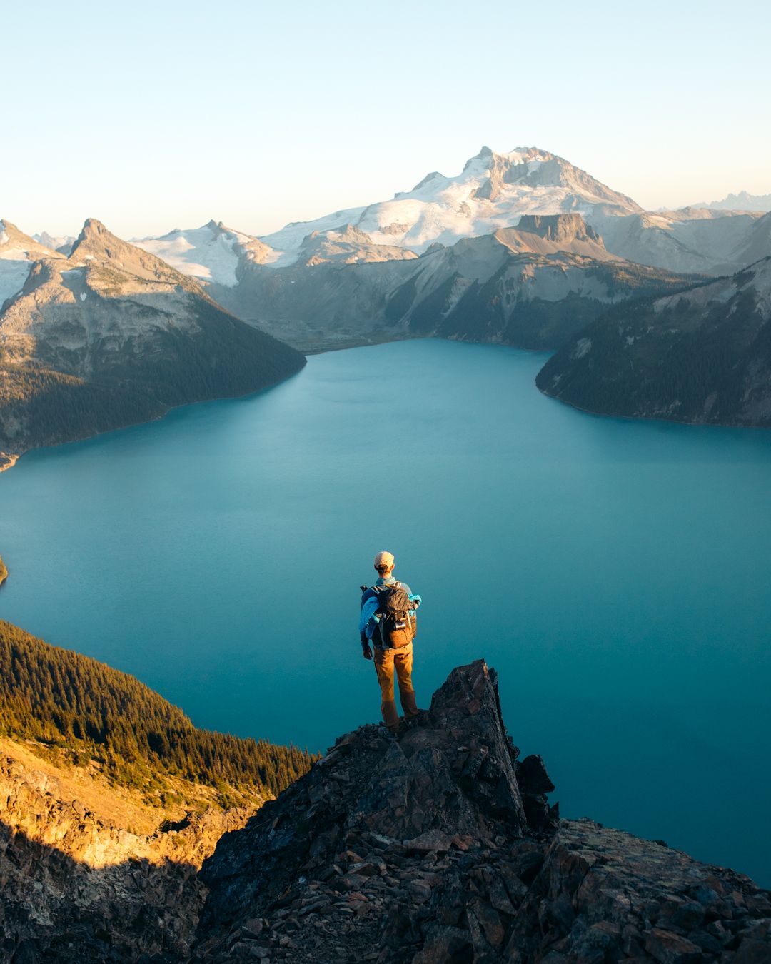 A hiker stands on a cliff edge overlooking a stunning turquoise mountain lake surrounded by peaks.
