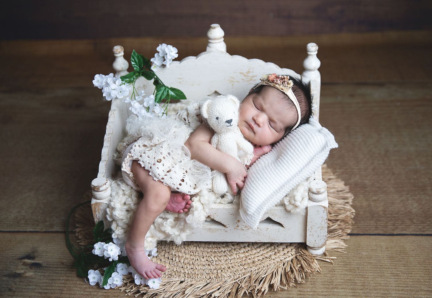 A tiny baby sleeps peacefully in a white vintage-style doll bed adorned with delicate flowers on a wooden floor. At photoshoot in Sidcup