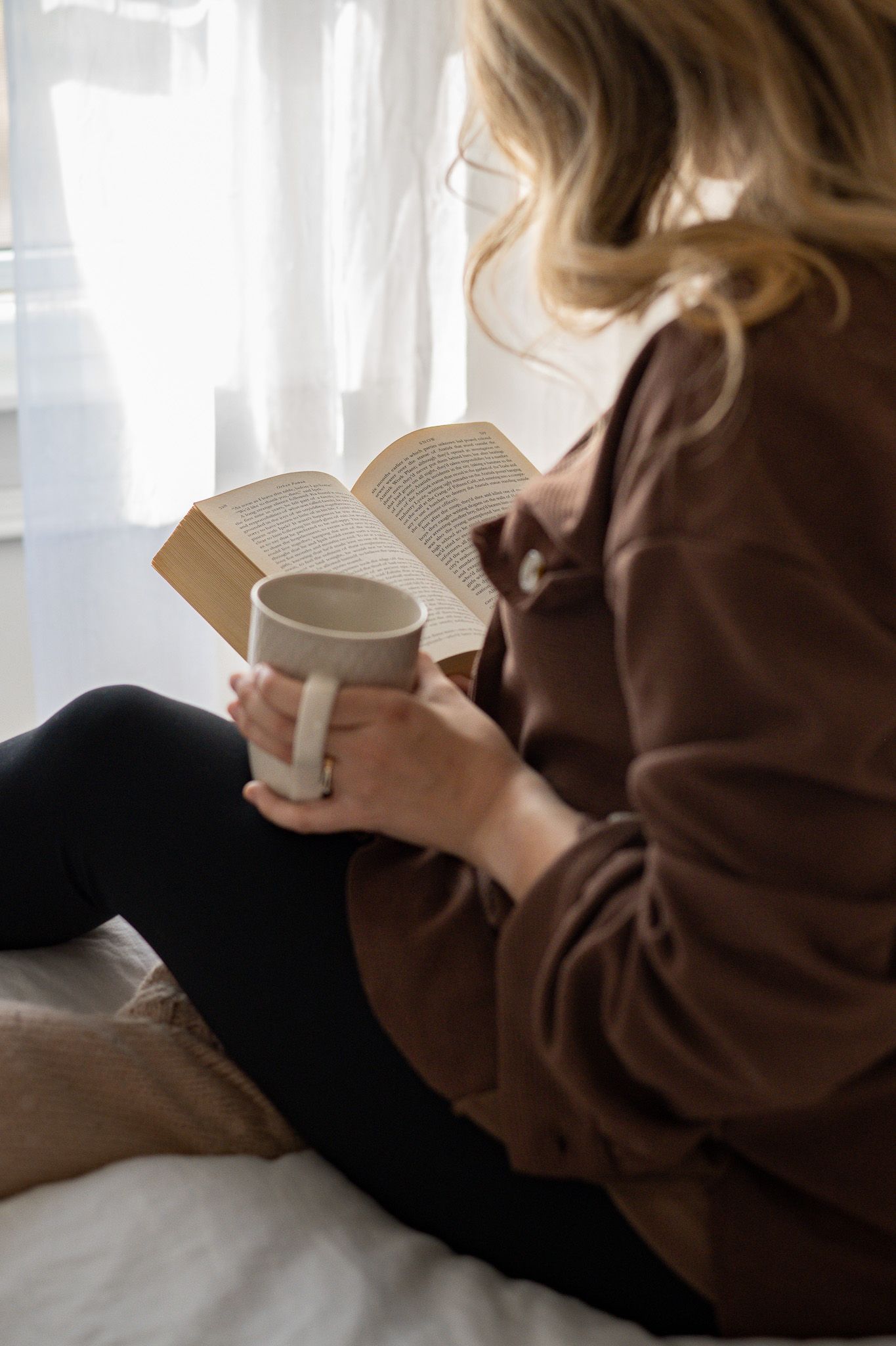 Cozy reading scene with brown sweater, book and white coffee mug by window with soft natural lighting.