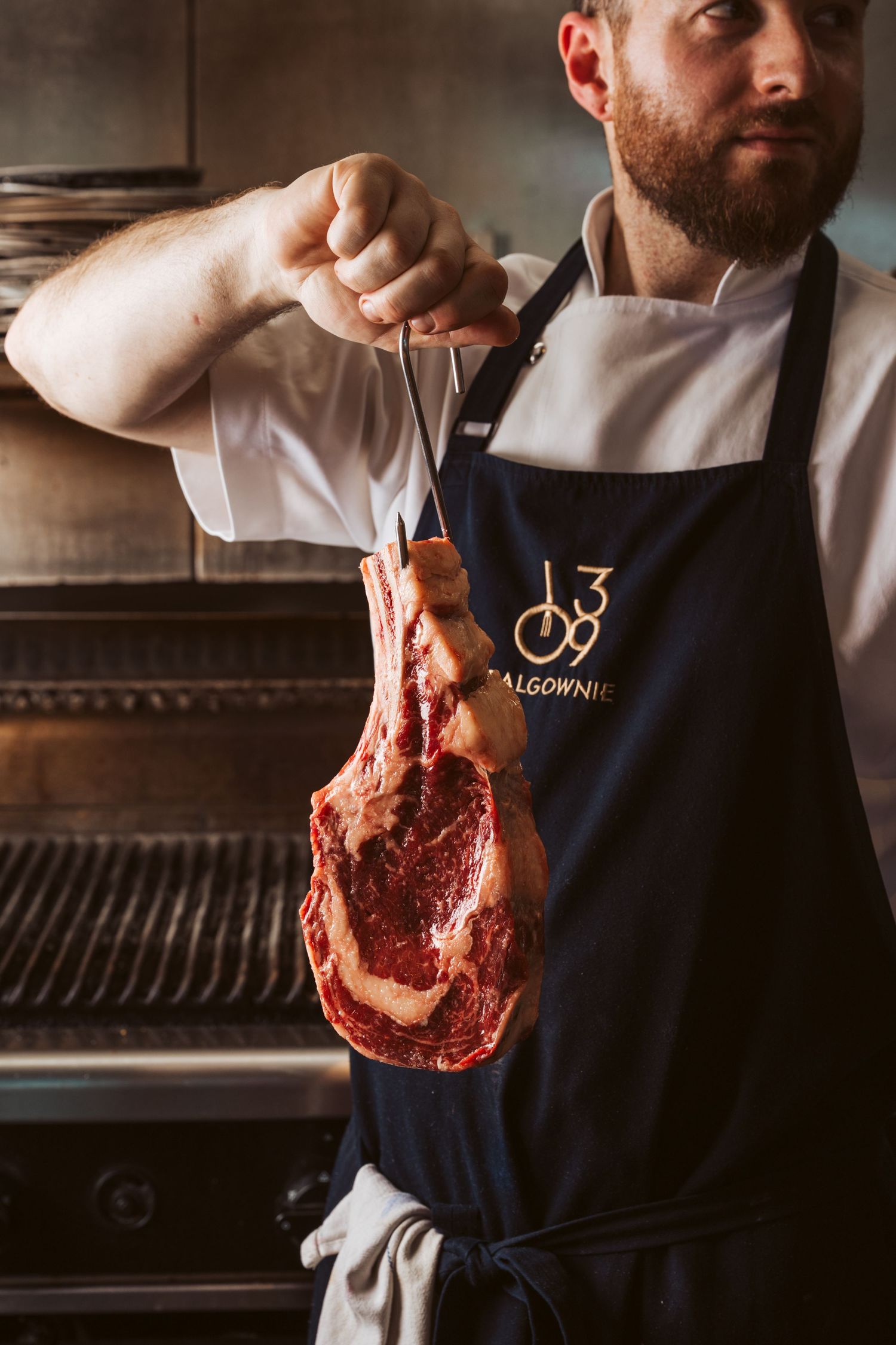 Hospitality branding photography featuring chef presenting premium dry-aged steak in commercial kitchen
