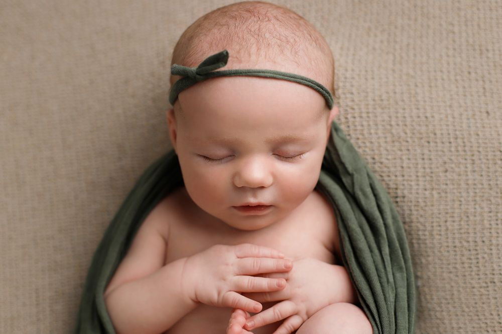 Peaceful sleeping newborn baby wrapped in olive green swaddle with matching headband resting on beige backdrop. Utah newborn photographer