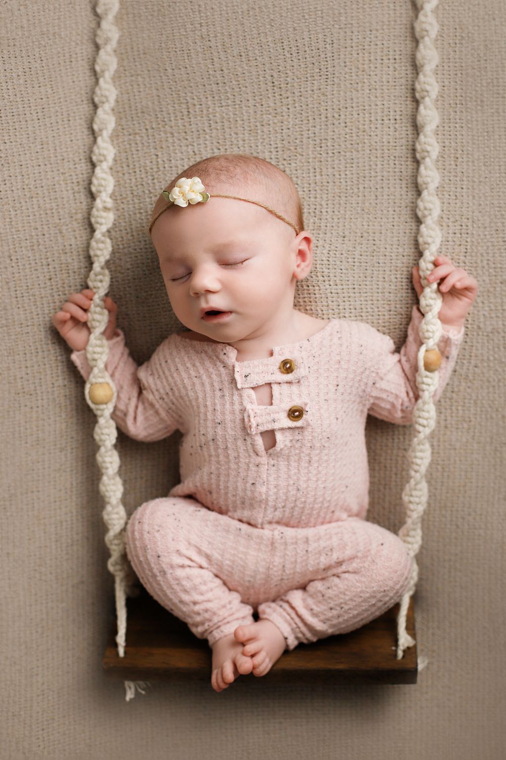 Tiny baby in pink knit outfit sleeps peacefully on a miniature wooden swing with macrame rope details. Utah newborn photography