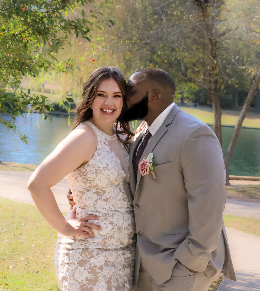 Newlywed couple poses outdoors by a lake in wedding attire on a sunny day.
