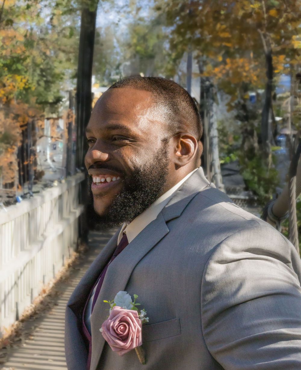 A cheerful groom in a grey suit with pink boutonniere poses on an autumn bridge.