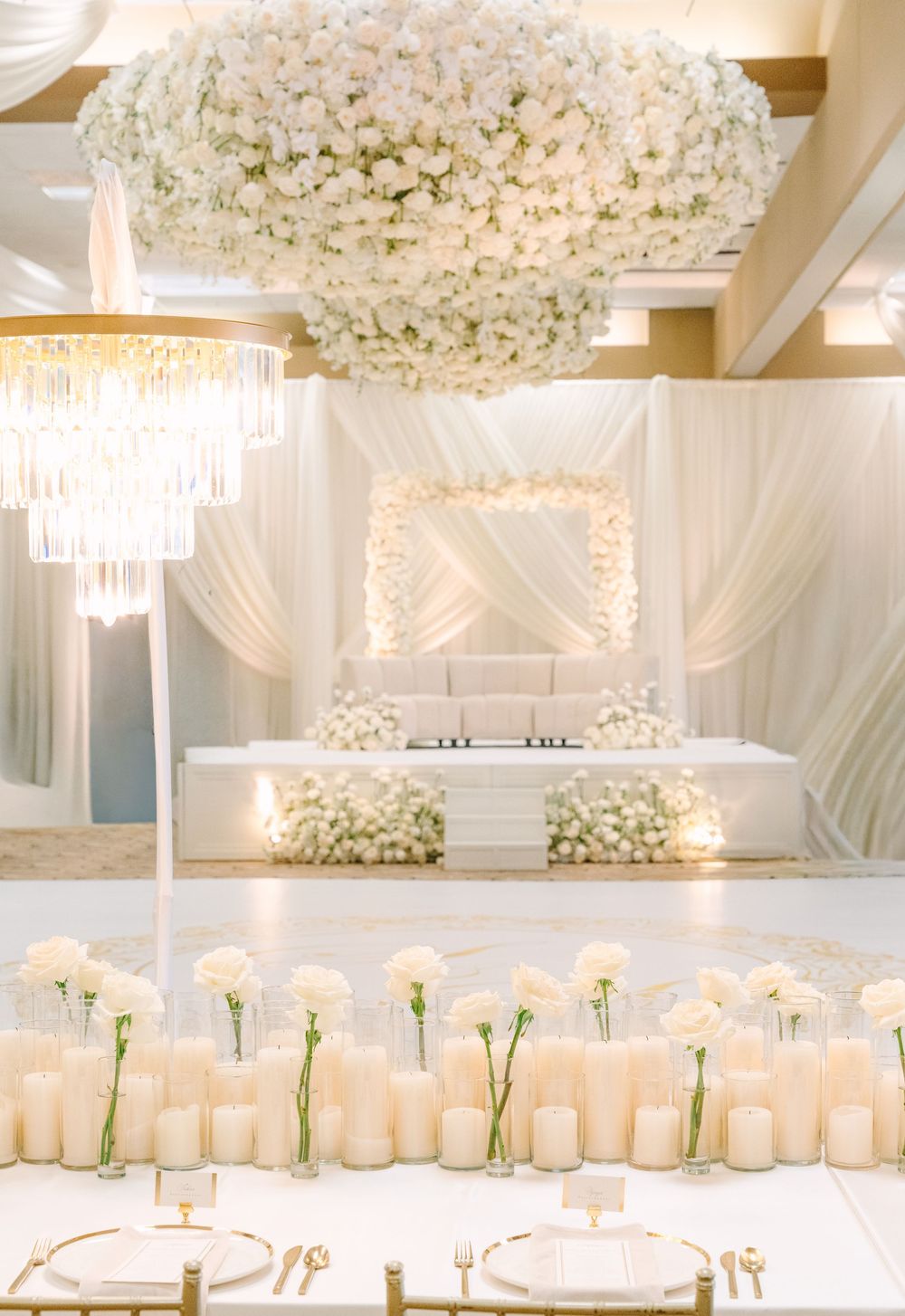 Elegant all-white wedding reception with floral chandelier, draped backdrop, candle centerpieces, and lush flower arrangements.