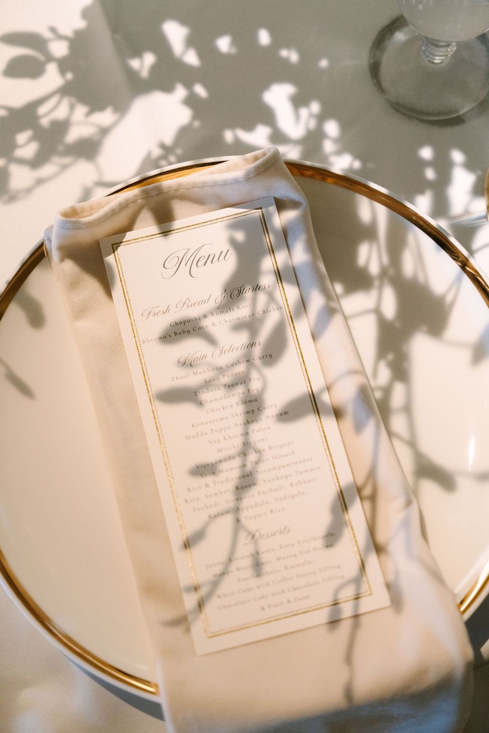 Elegant wedding menu card on gold-rimmed plate with floral shadows, white napkin, and glass in soft natural light.