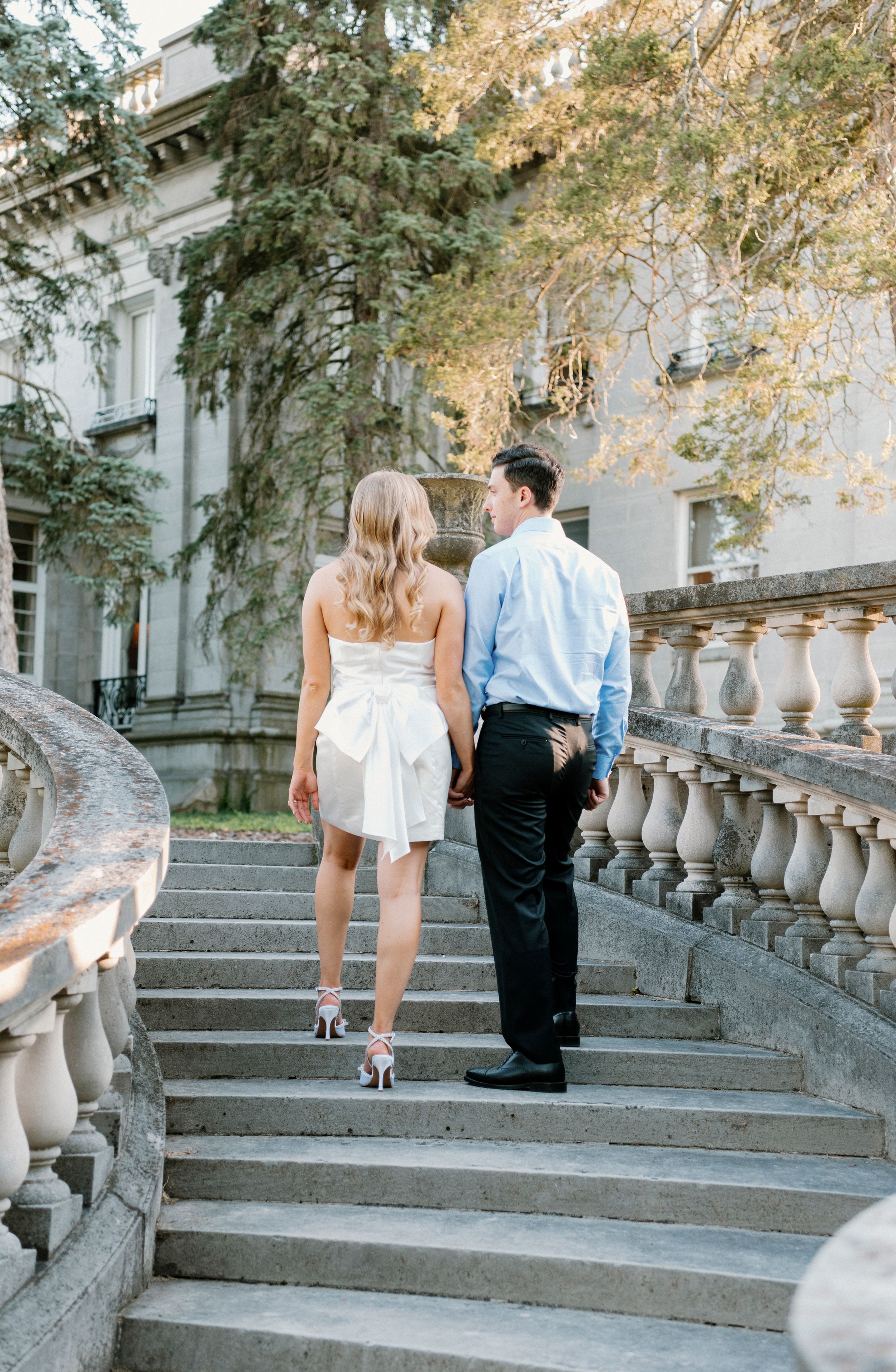 A couple walks up elegant stone stairs with ornate balustrades at a historic mansion during a romantic summer afternoon.