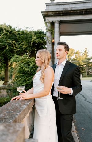 A couple in wedding attire holds champagne glasses while standing on an outdoor terrace with classical columns.