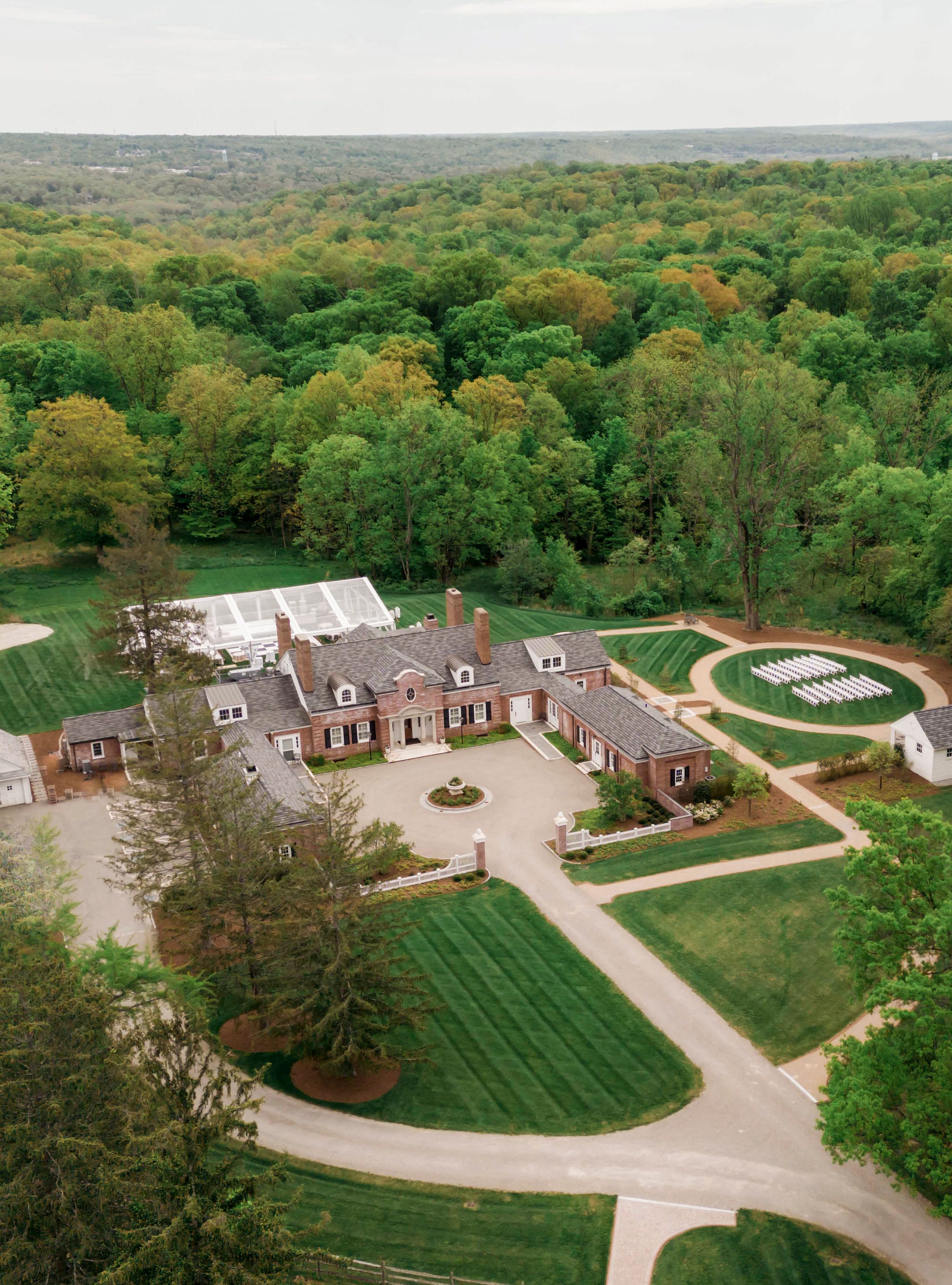 Aerial view of an elegant brick mansion estate with curved driveway and manicured grounds surrounded by lush green forest.