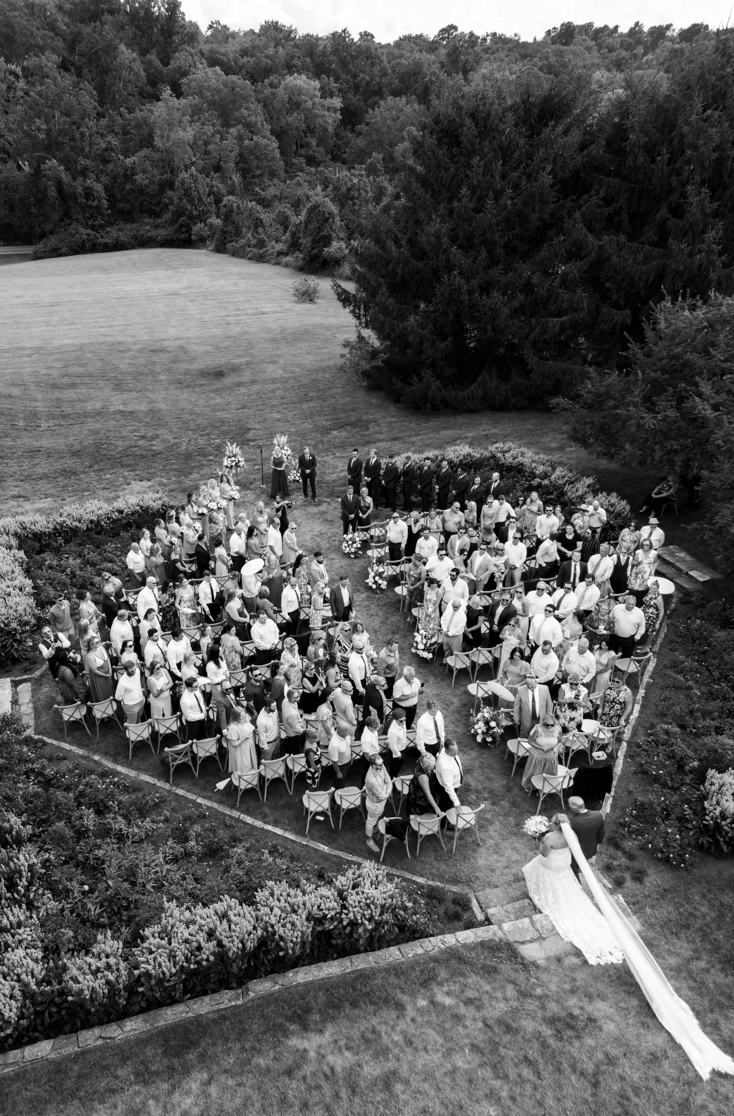Aerial view of a large outdoor wedding ceremony with guests gathered on a grassy lawn surrounded by trees.