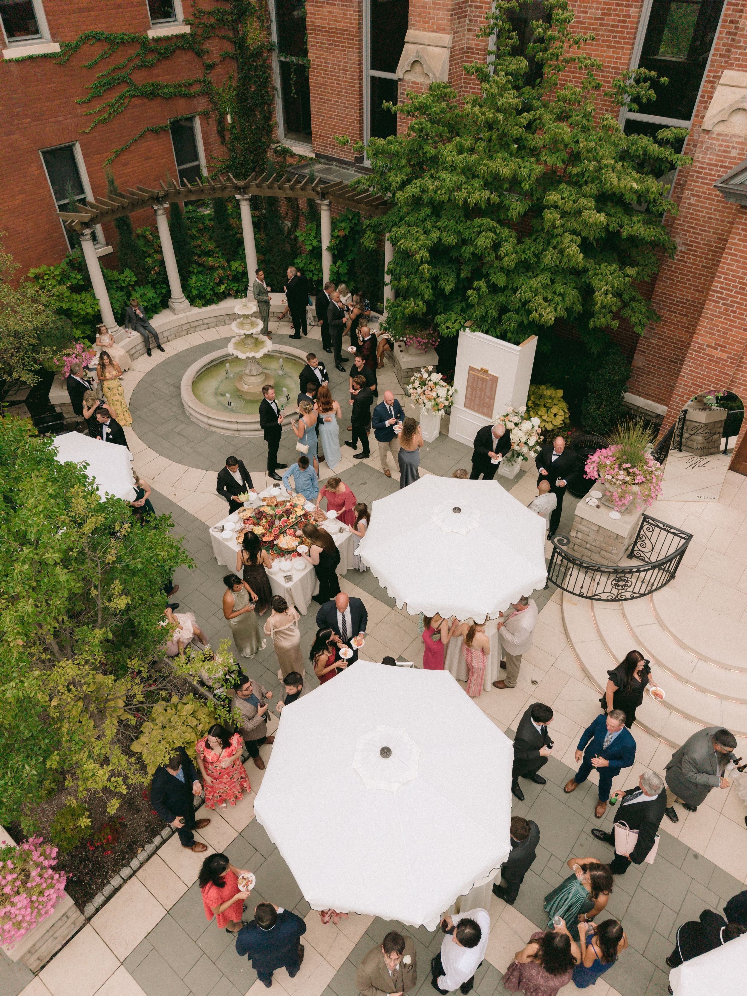 Aerial view of an elegant outdoor wedding reception in a brick courtyard with white umbrellas and a fountain.