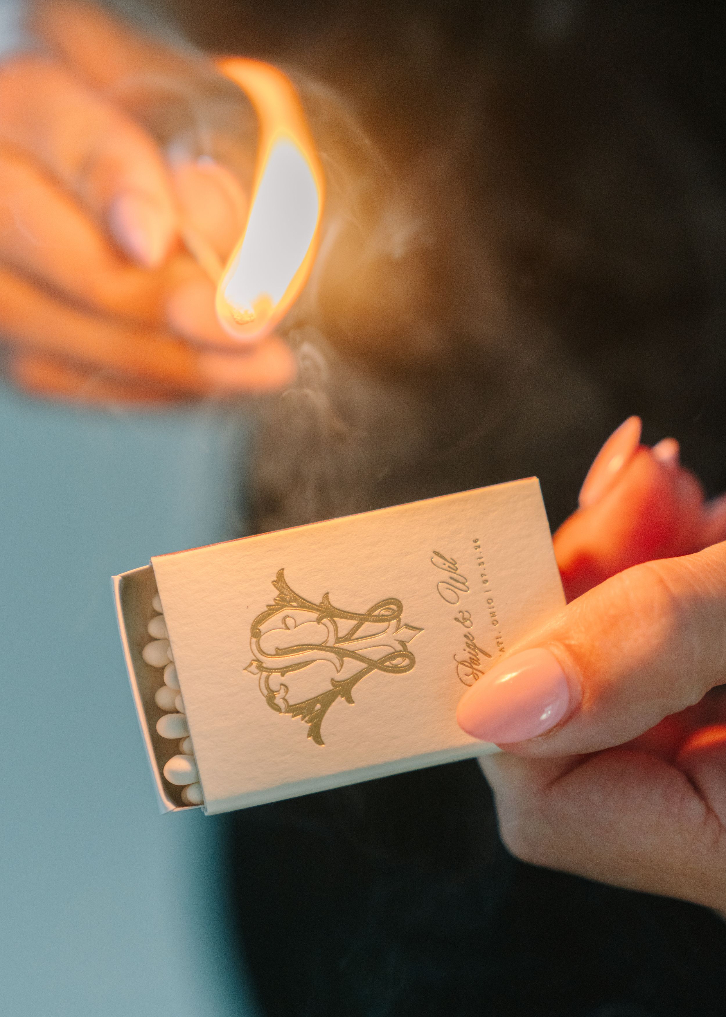 Hands holding an elegant gold monogram matchbox with a lit match producing smoke above it.