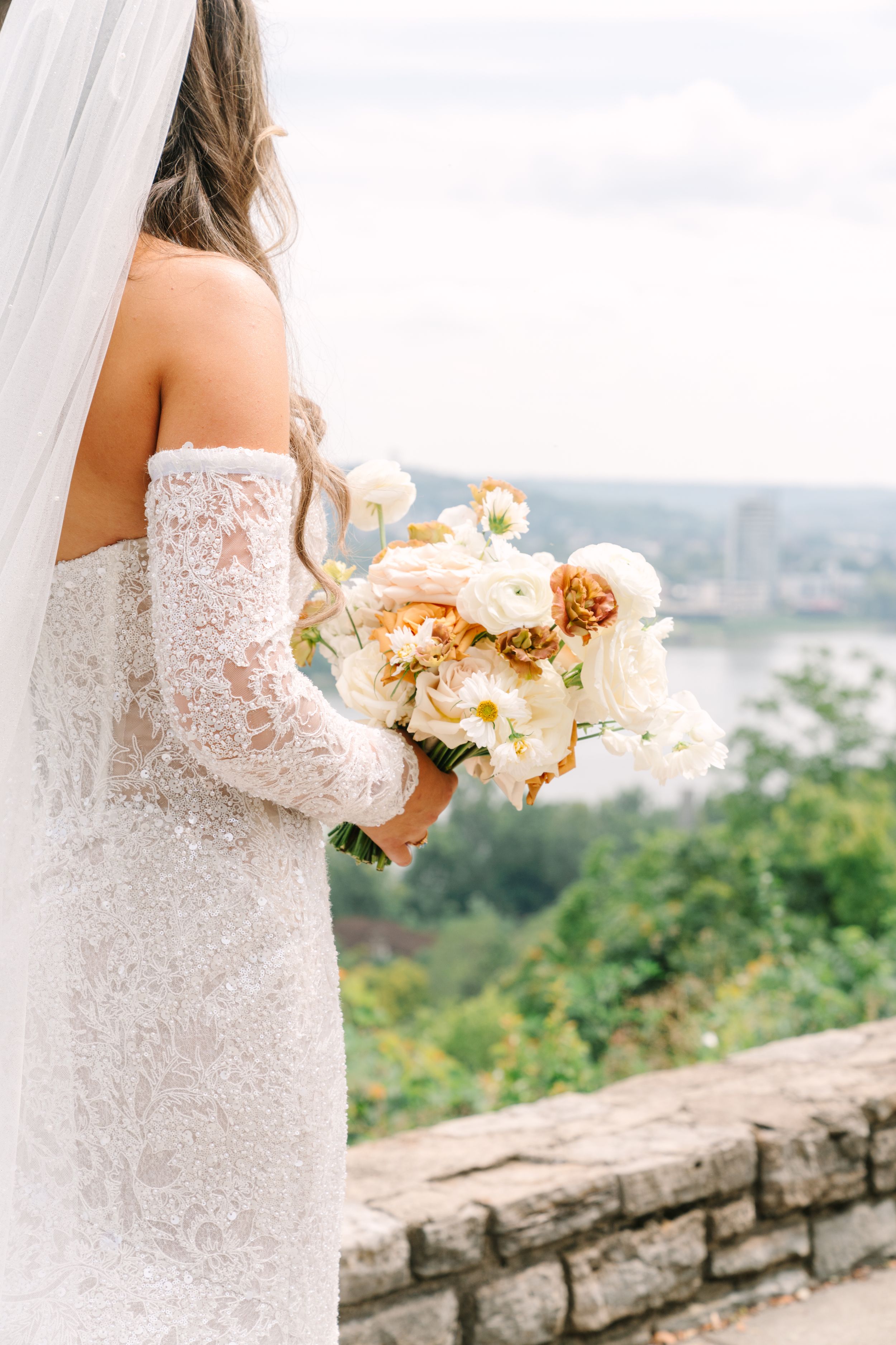 Bride in lace off-shoulder gown holding peach and white floral bouquet, with scenic river view backdrop.