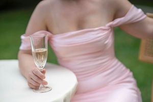 Someone in a pink dress holds a champagne flute while sitting at a white table outdoors.