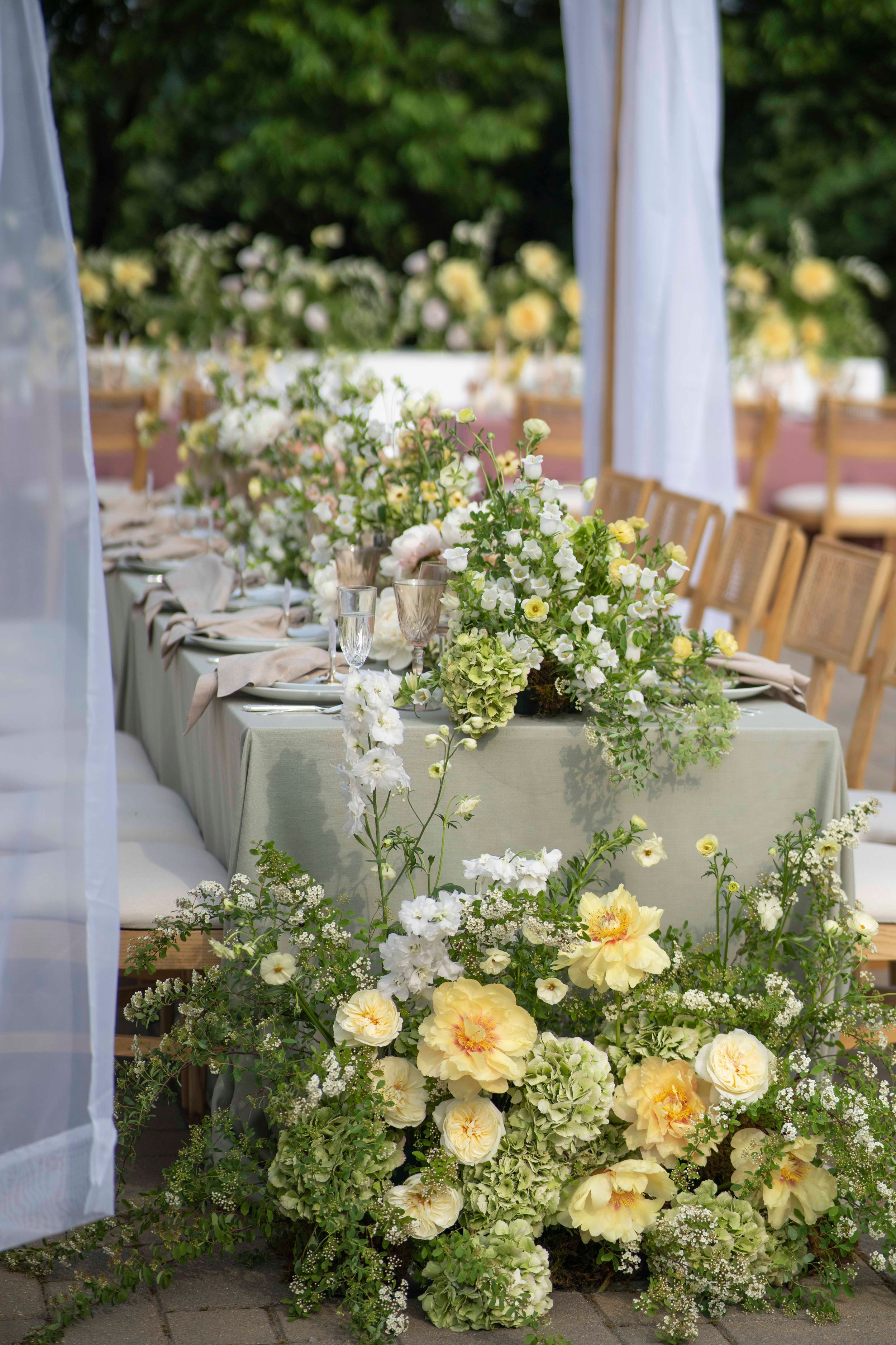 Elegant outdoor wedding table with neutral tablecloth and lush floral arrangements of pale yellow roses and white wildflowers.