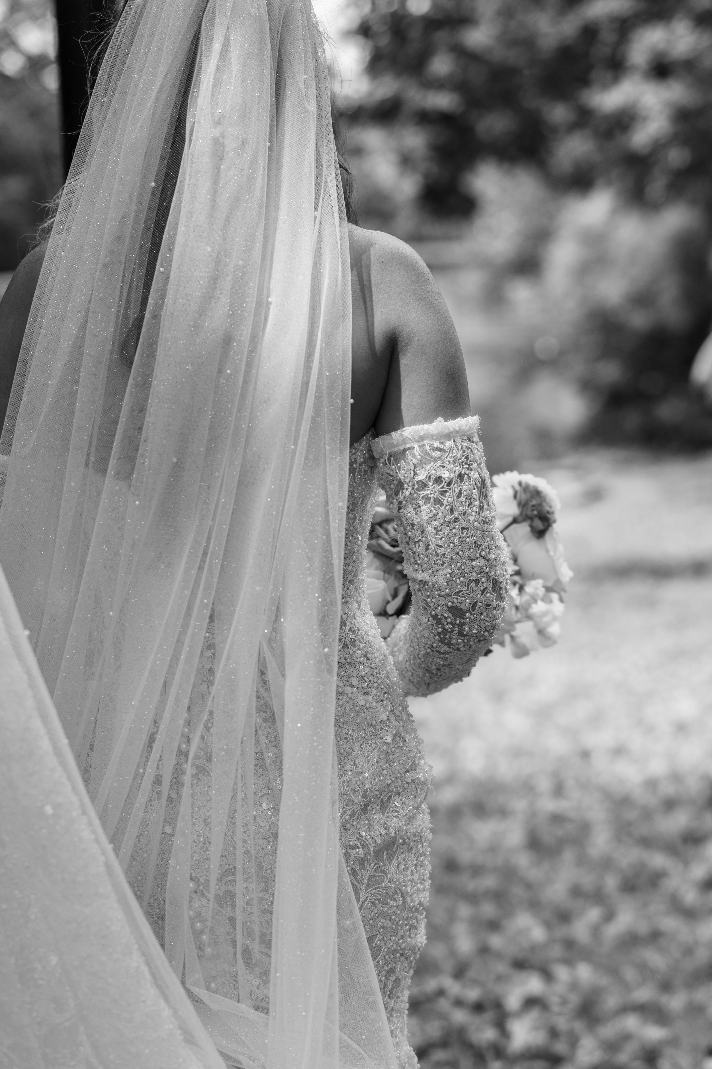 Black and white photo of a bride in lace gown holding bouquet, partially hidden behind flowing wedding veil outdoors.