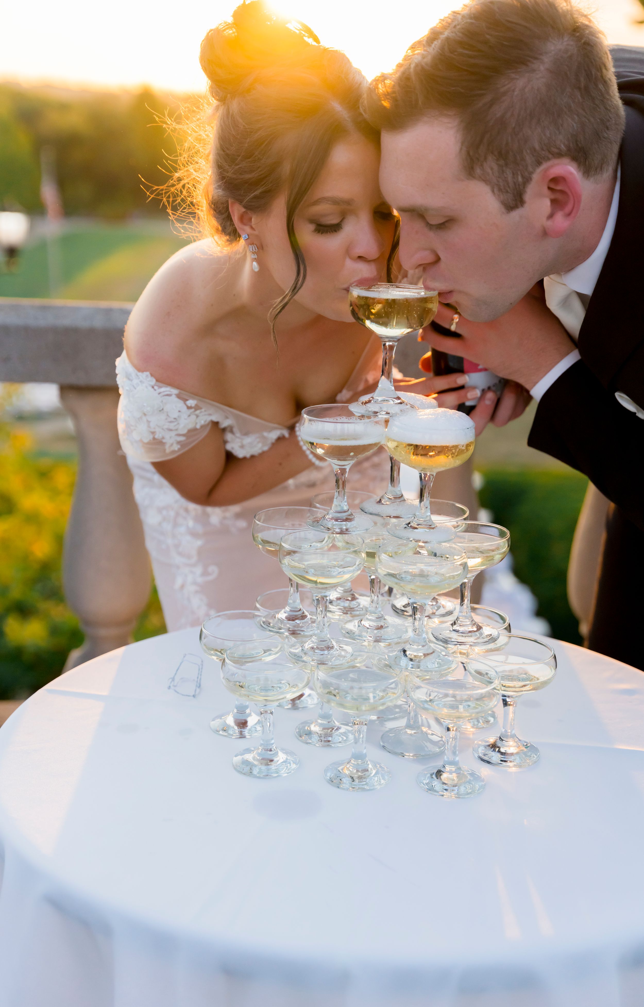 Couple sips champagne together from a tower of crystal glasses at sunset during elegant wedding celebration.