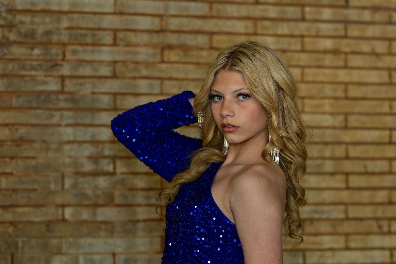 Model in sparkly blue dress posing against brick wall background.