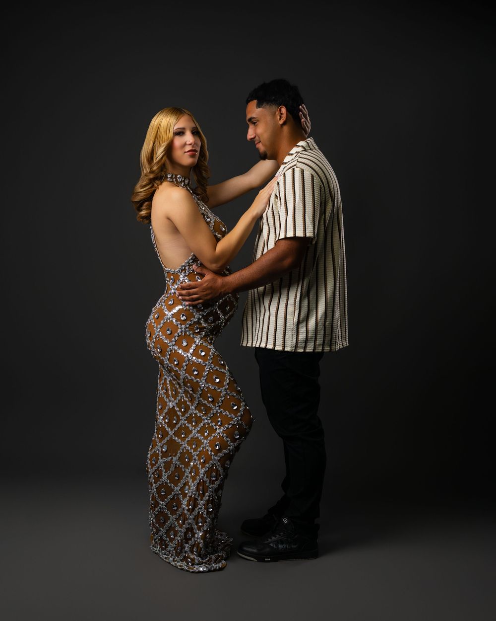 A couple poses in a close embrace in a studio, she in a sparkling beaded halter gown and he in a striped short-sleeve shirt.