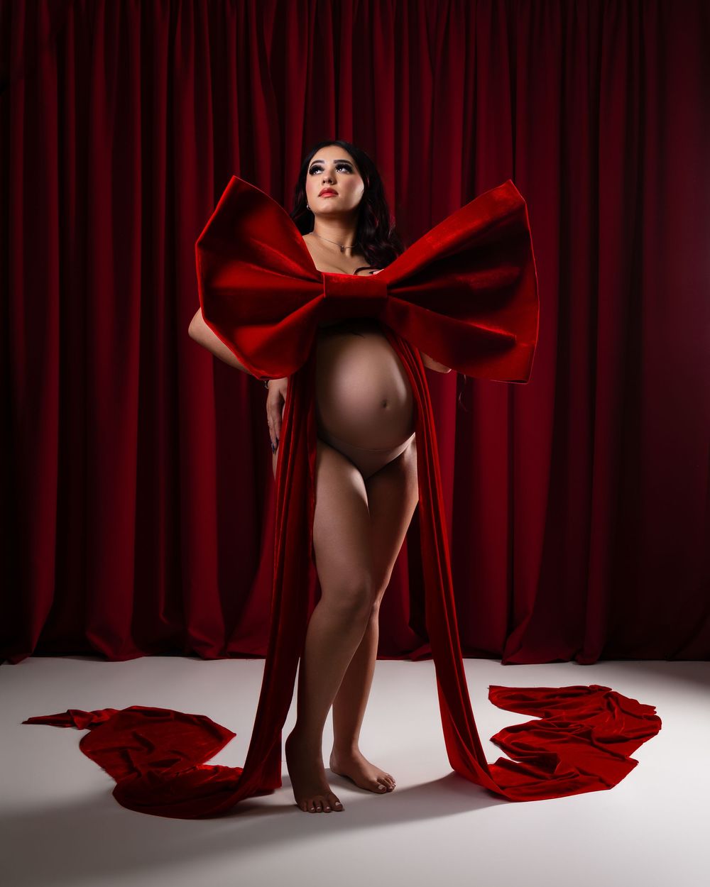 Pregnant woman poses in a maternity photoshoot wrapped in a giant red velvet bow against a deep red curtain backdrop.