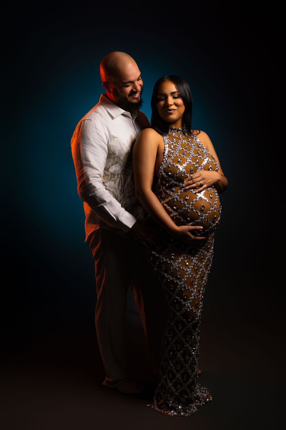Smiling expectant couple in a studio maternity portrait, the woman in a beaded gown cradling her bump as her partner embraces her.