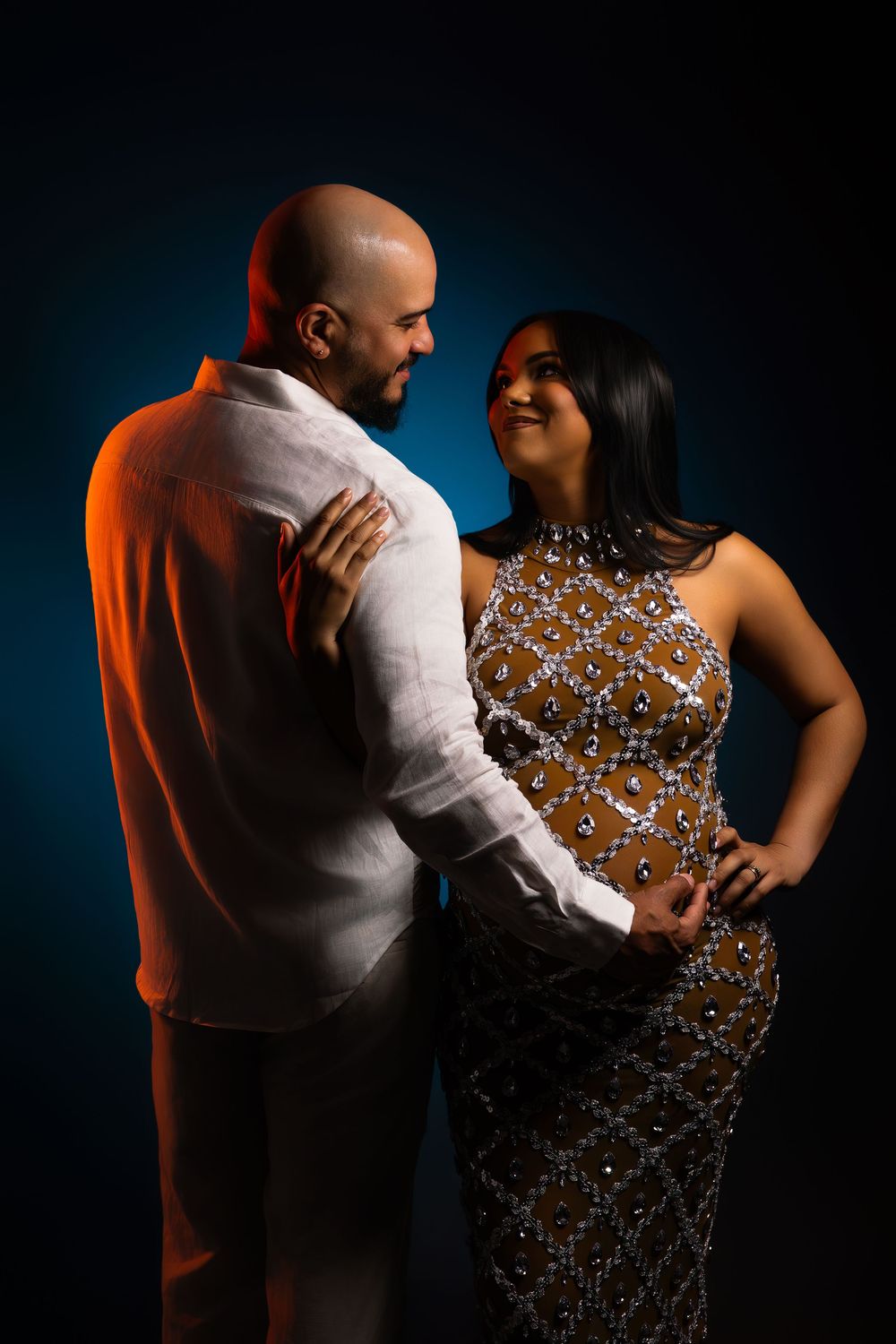 Smiling couple in a studio portrait, a bald bearded man in a white linen shirt embracing a woman in a sparkling beaded gown against a blue b
