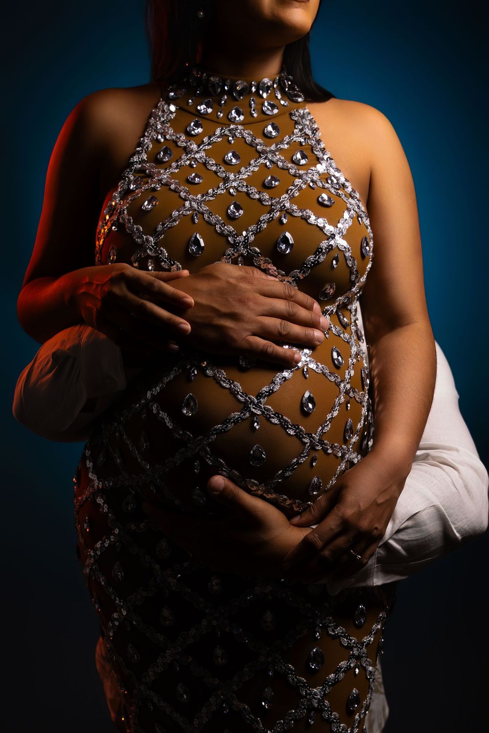 Pregnant woman in a crystal-embellished halter gown cradling her baby bump in dramatic blue and orange studio lighting.
