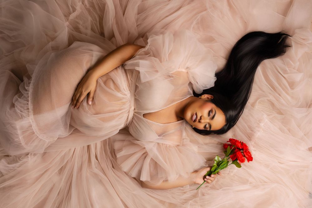 Pregnant woman with long black hair lying on a blush tulle gown, holding a red rose in a dreamy maternity photoshoot.