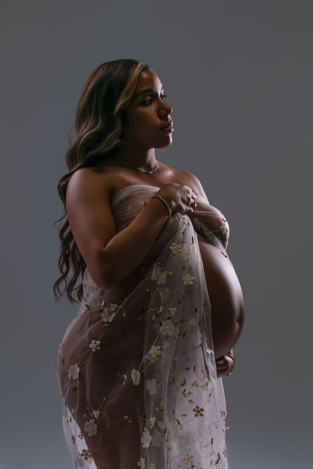 Pregnant woman in profile draped in sheer floral embroidered tulle, cradling her baby bump in a moody studio maternity portrait.