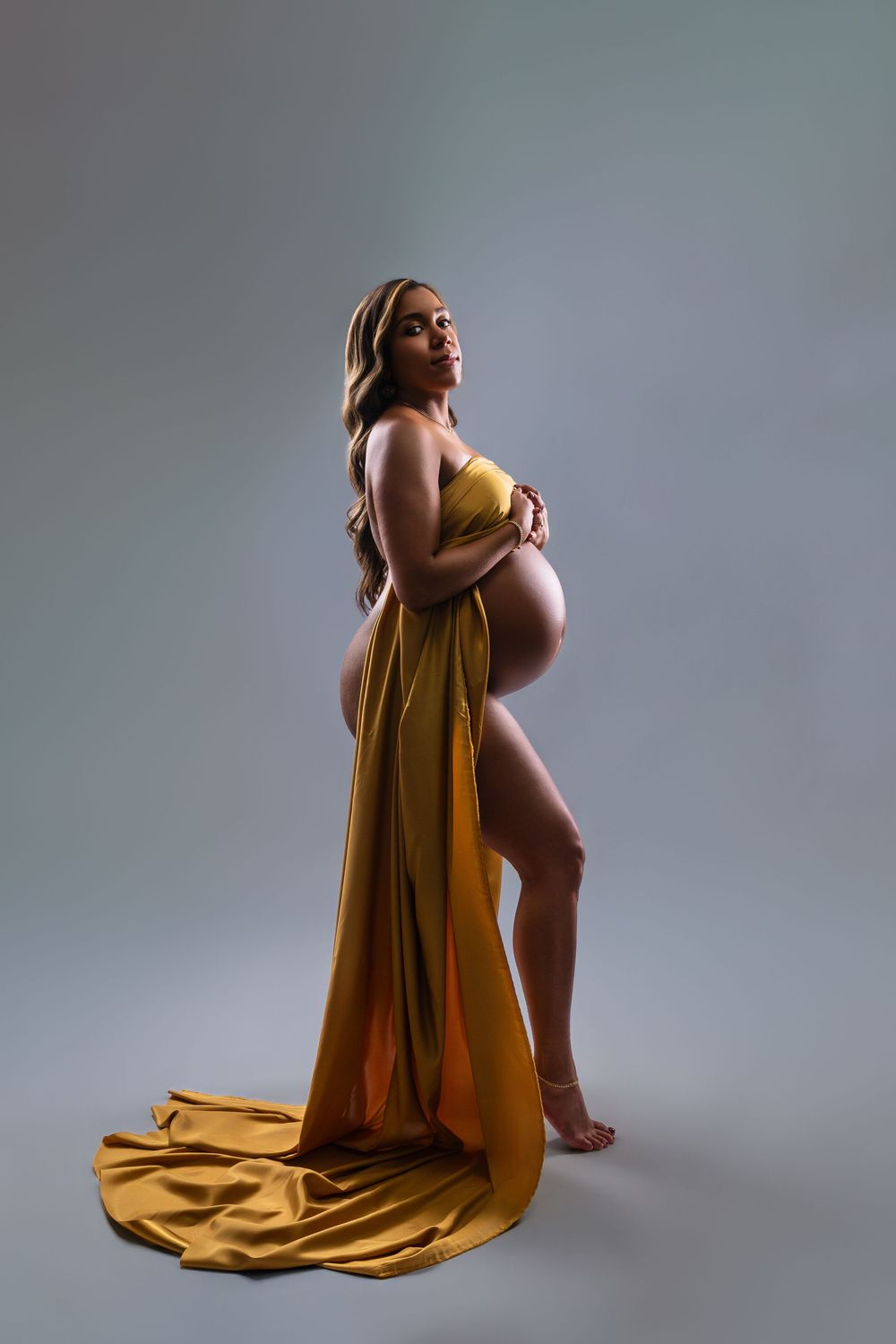 Pregnant woman posing in a studio maternity photoshoot, draped in flowing golden silk fabric with a long train against a gray backdrop.