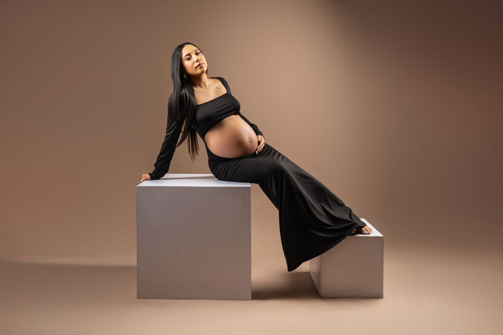 Pregnant woman in a black two-piece maternity outfit with flowing wide-leg pants poses on gray cubes in a beige studio maternity photoshoot.