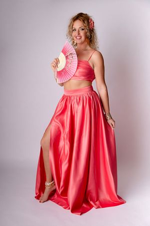 Une personne pose dans une robe deux pièces rose corail avec une fente, tenant un éventail décoratif, sur un fond gris clair.
