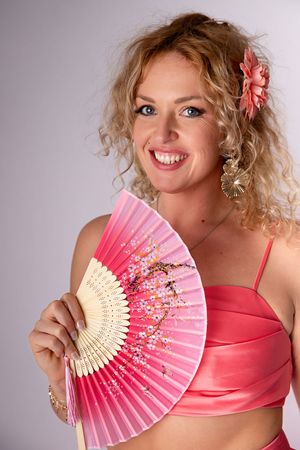 Une femme souriante aux cheveux blonds et bouclés tient un éventail décoratif rose tout en portant un haut couleur corail et un accessoire