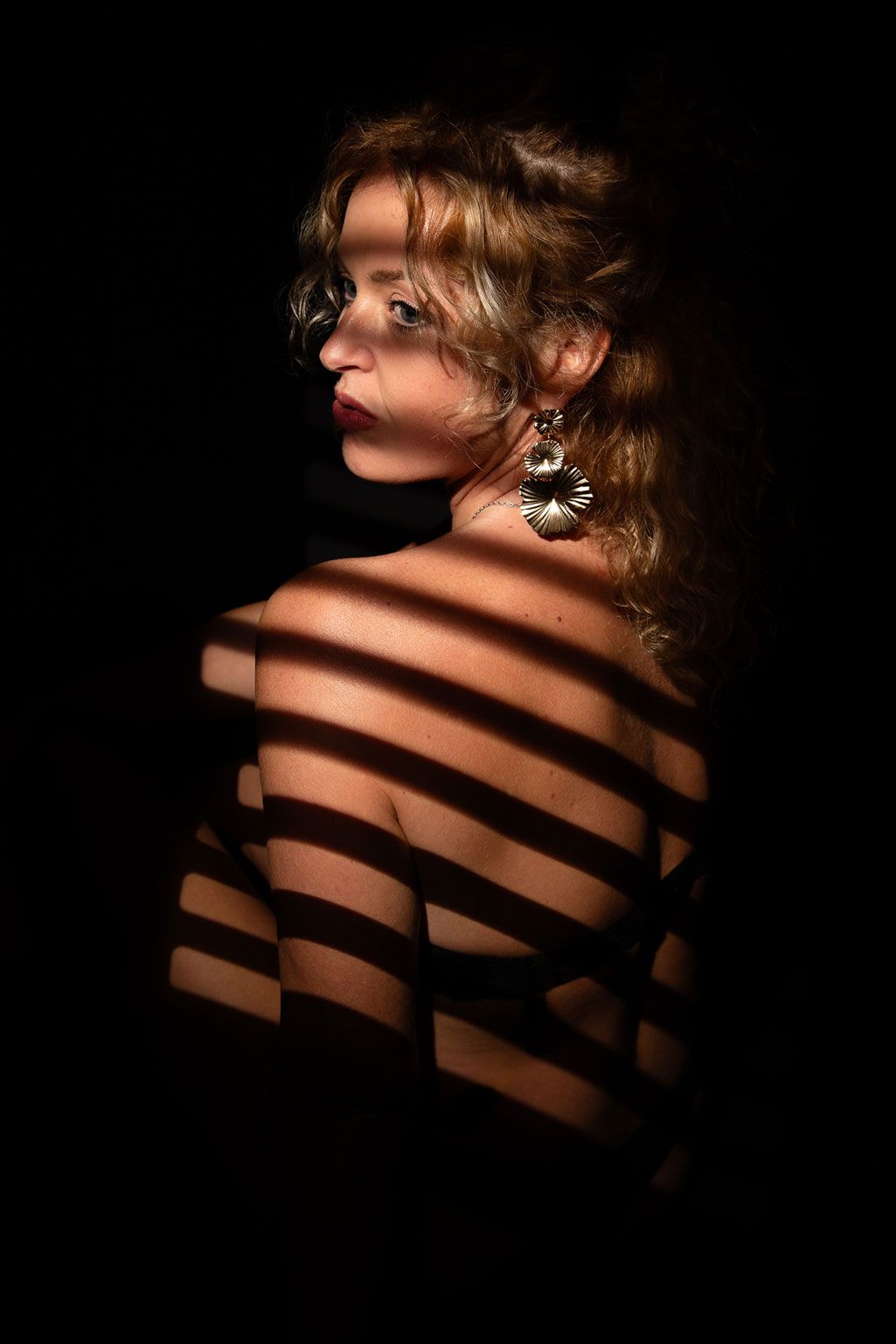 Portrait dramatique avec des ombres rayées tombant sur des cheveux blonds bouclés et des épaules nues, sur un fond sombre.