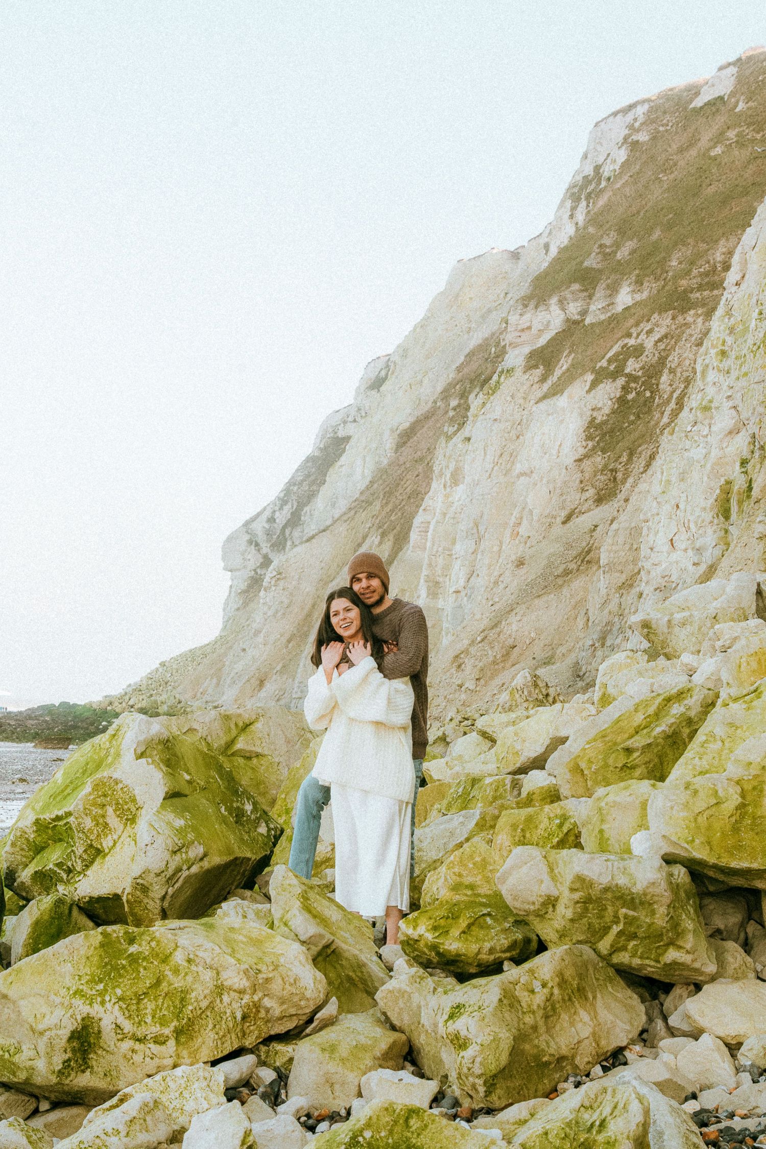 Two people embracing on mossy rocks beneath towering white cliffs along a coastal shore.