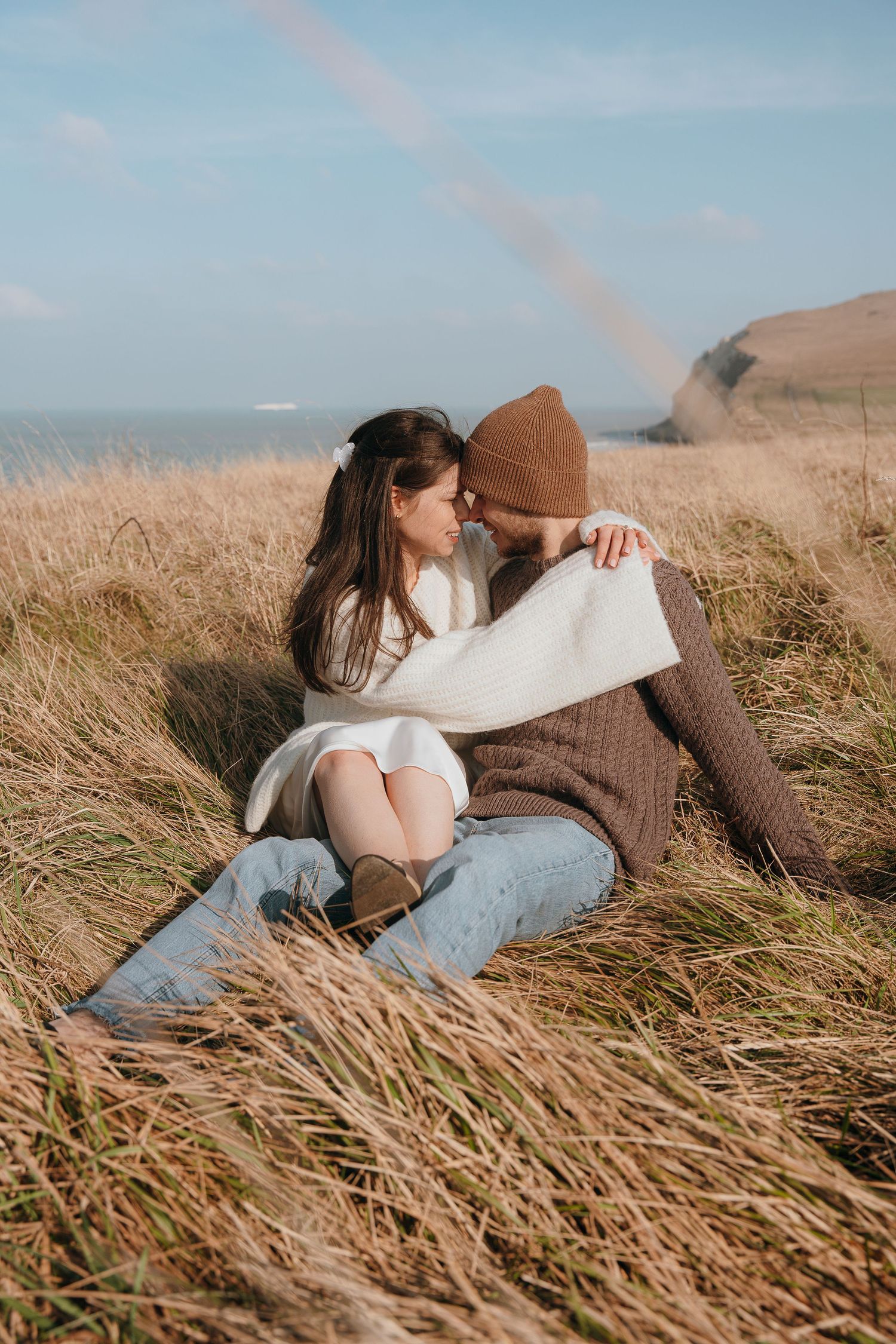 A couple sits together in tall grass on a hillside overlooking the ocean during late afternoon sunlight.