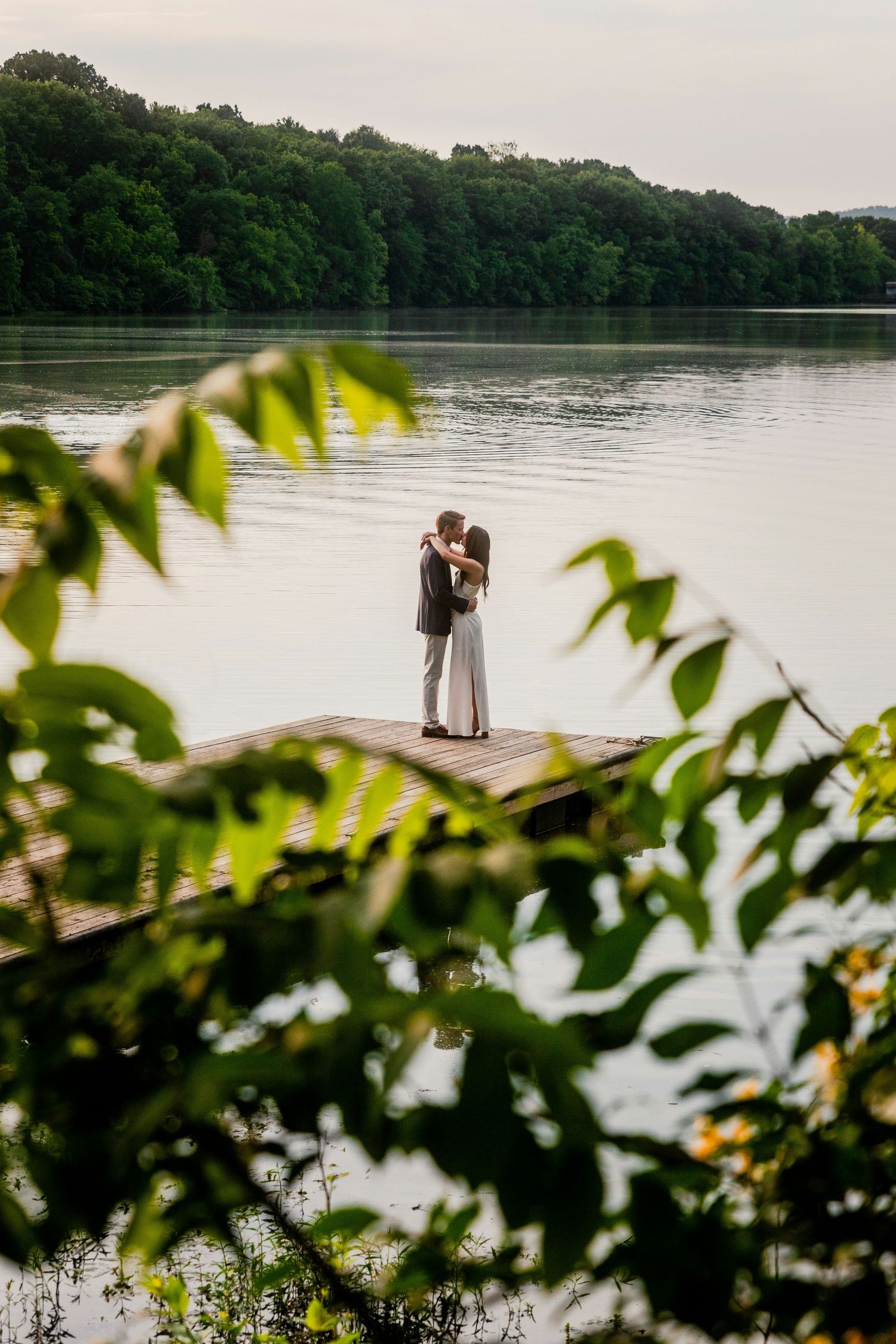 Two figures embrace on a wooden dock overlooking a serene lake surrounded by lush green forest.