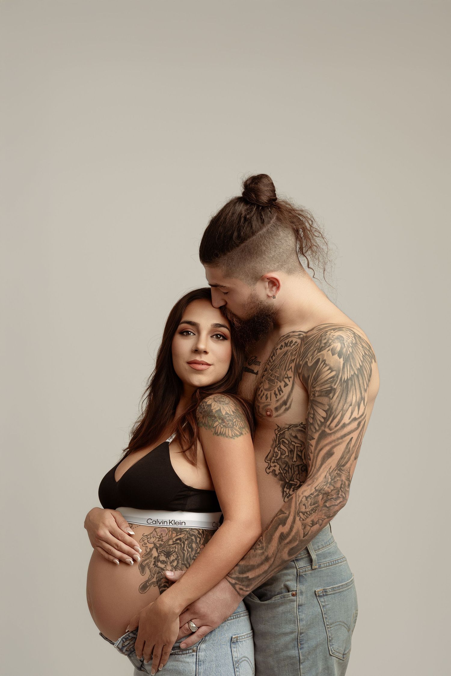 Tender maternity portrait of couple in dark clothing against brown studio background.