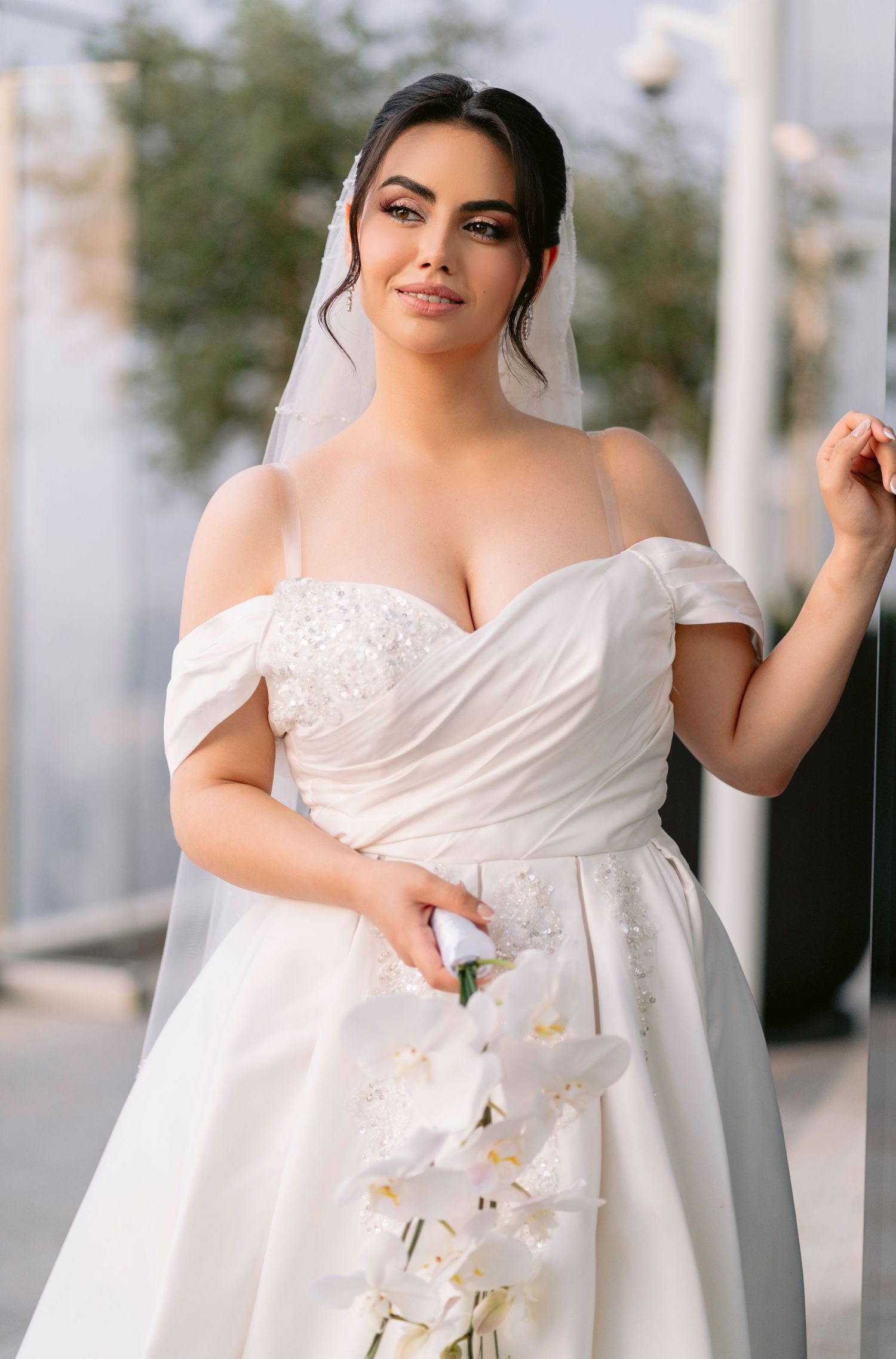 A bride in a classic white wedding dress poses elegantly with white orchids in an outdoor setting.