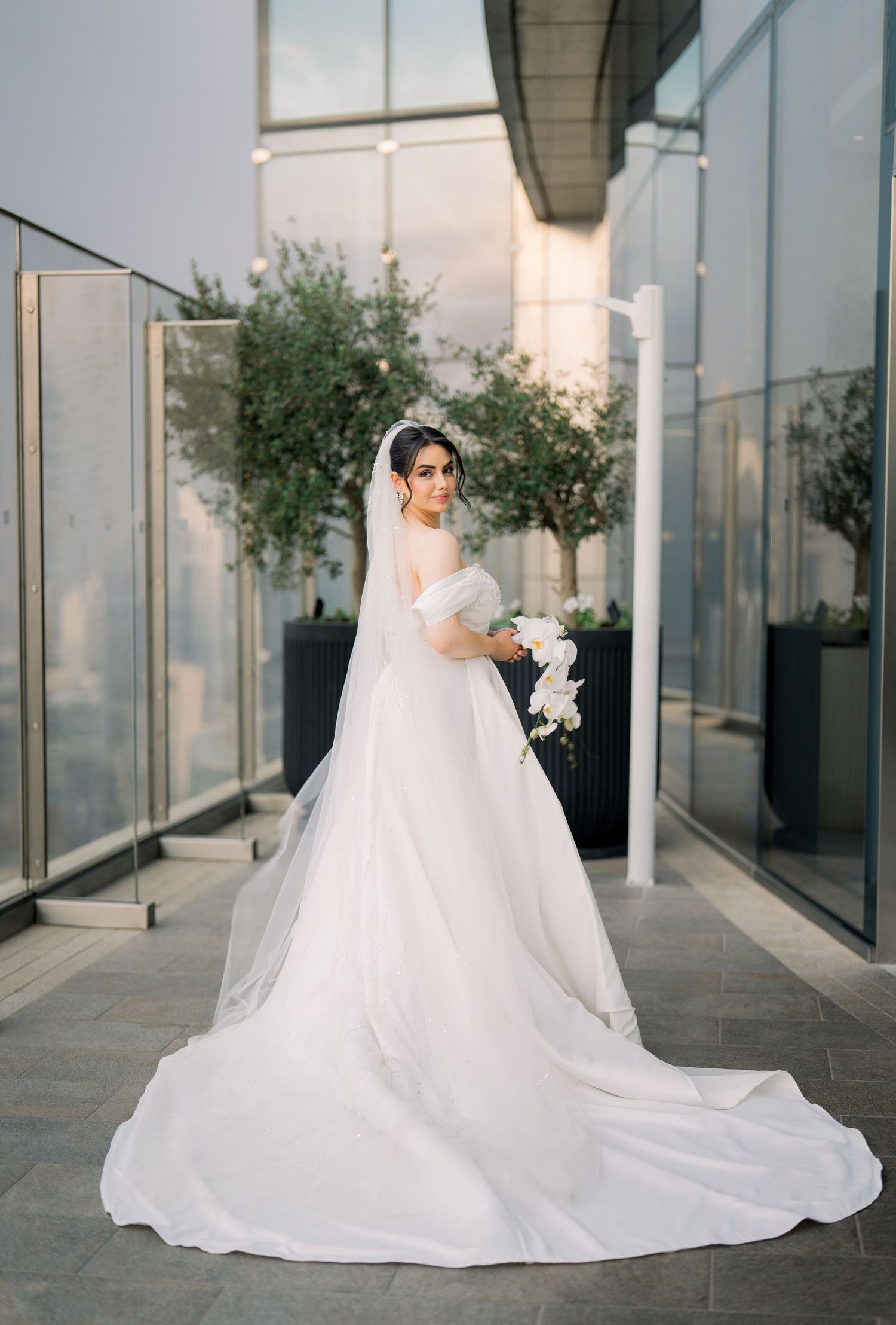 Stunning bride in a classic white ball gown with cathedral train stands on a modern terrace with city views.