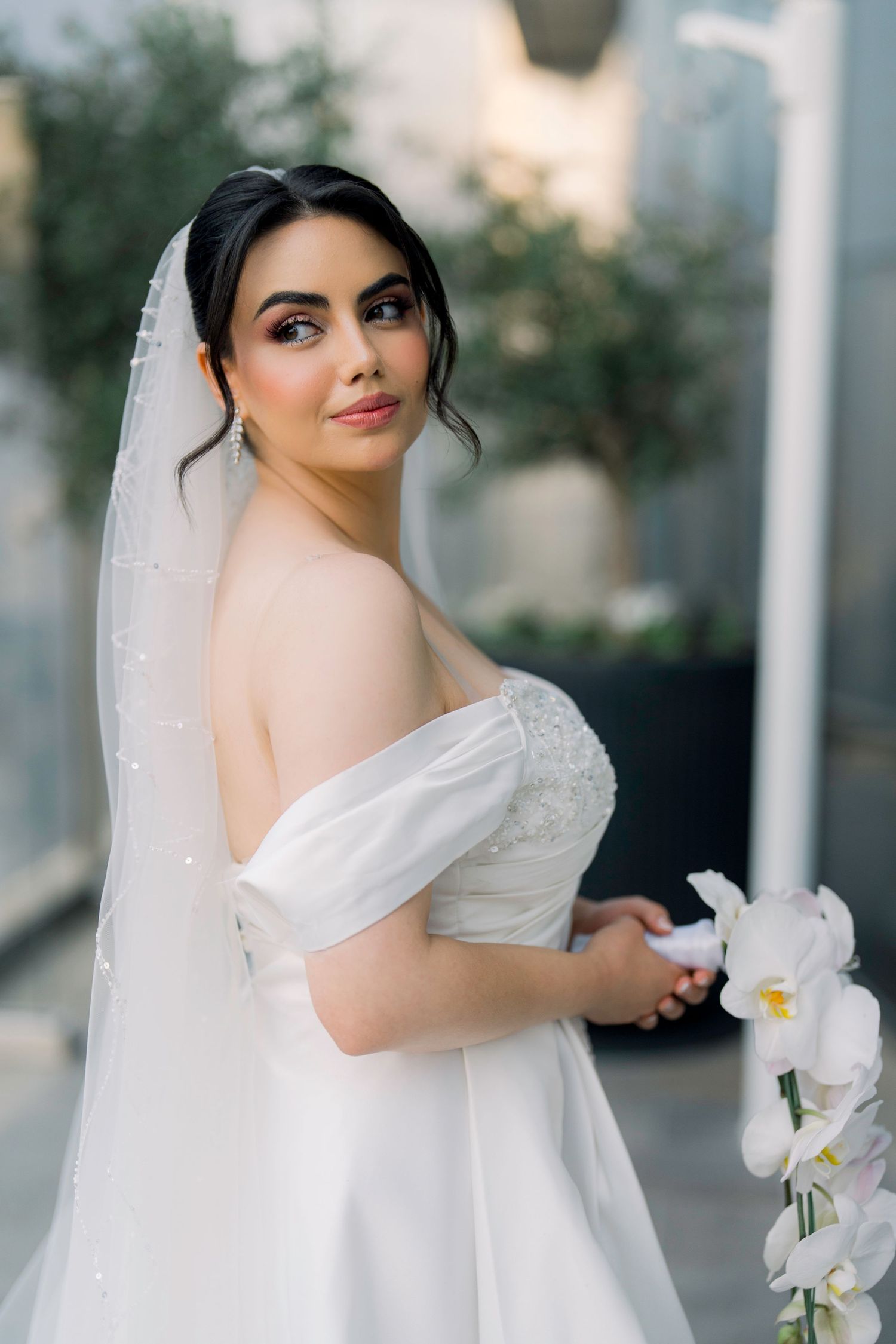 Elegant bride in a white off-shoulder gown with a flowing veil poses with white orchids against a modern backdrop.