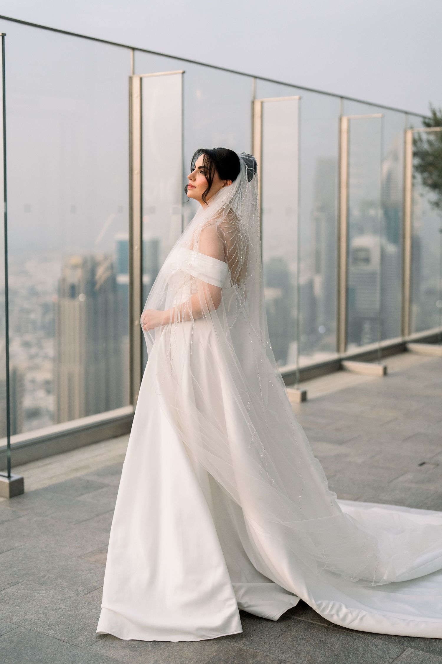 A bride in a classic white wedding dress and veil poses on a rooftop terrace with city views in the background.