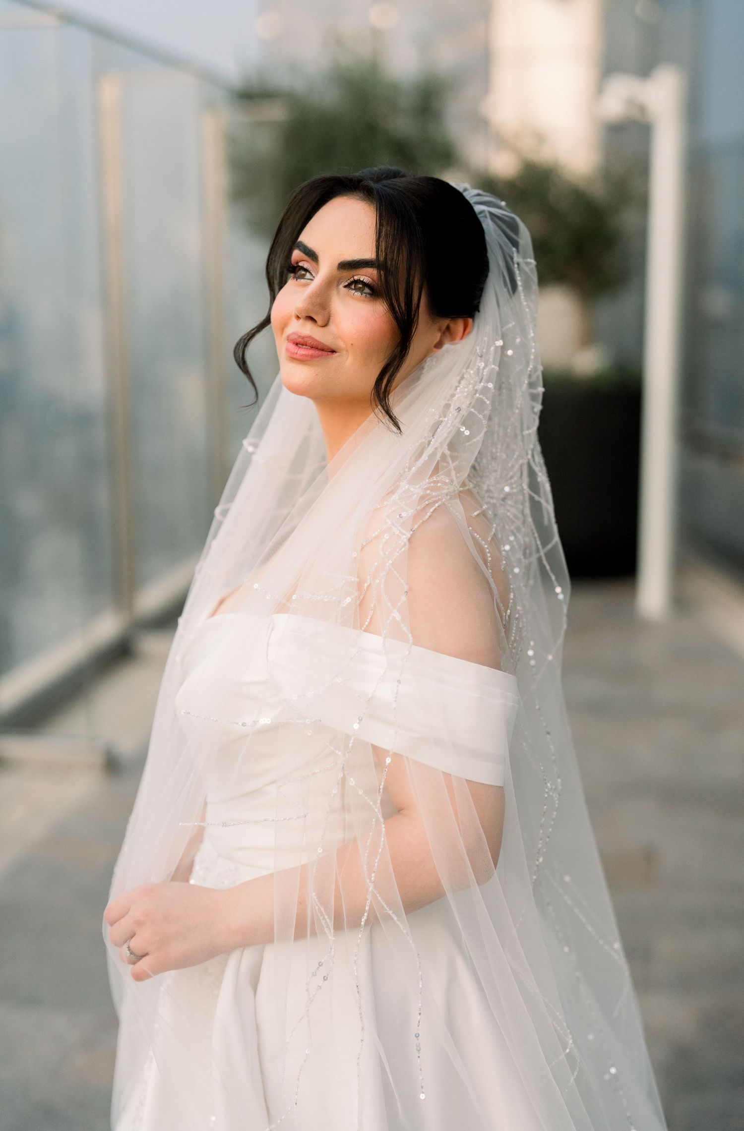 A bride in a white off-shoulder wedding dress poses elegantly with a flowing veil in a modern architectural setting.