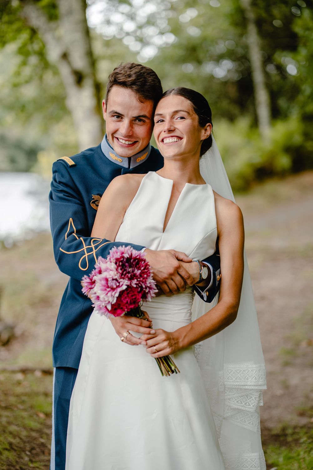 A couple in wedding attire poses with a pink bouquet in an outdoor setting.