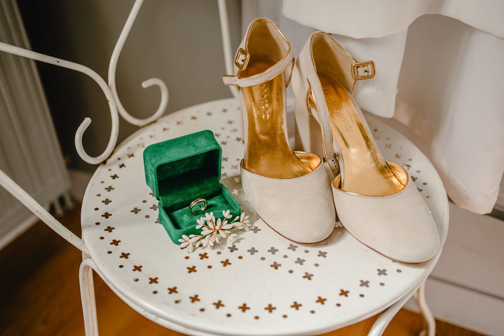 White bridal shoes and emerald green ring box displayed on decorative white metal table.