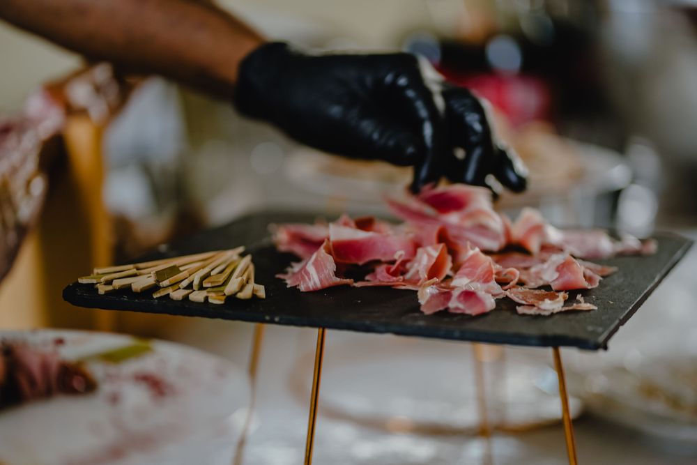 Chef preparing meat appetizers on dark slate board wearing black gloves.