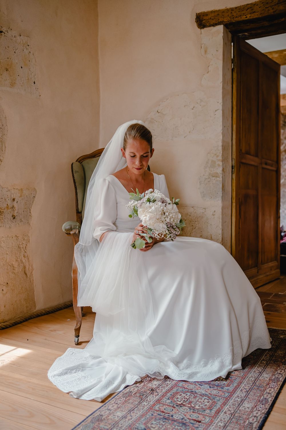 Bride in white dress sitting on patterned rug in rustic room with natural light.