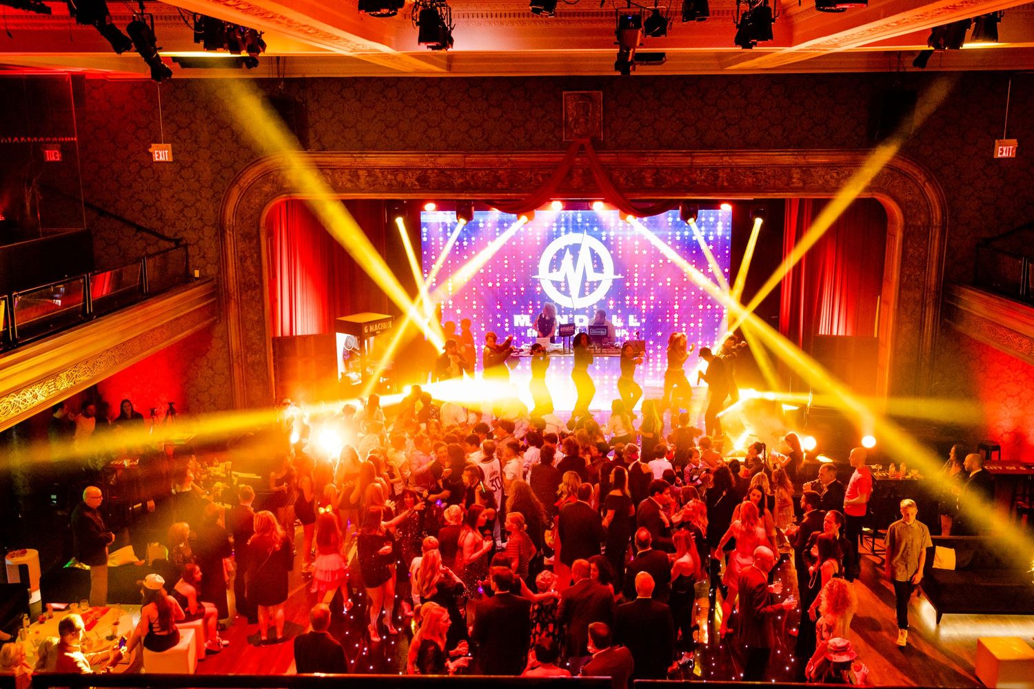 Concert venue lit with dramatic orange and yellow spotlights over a crowded dance floor.