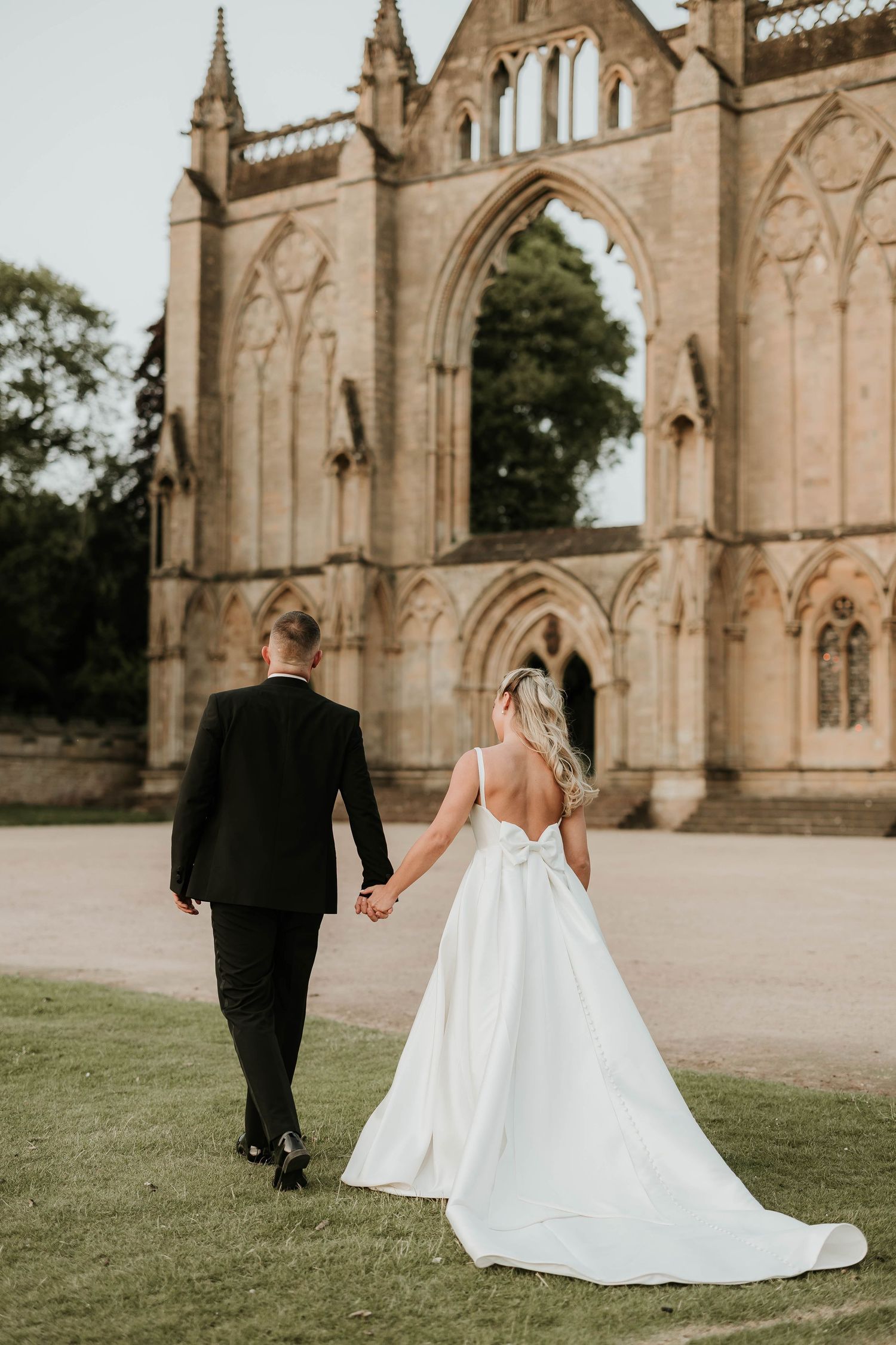 Bride and groom walk along the grounds of a Gothic cathedral with ornate stone architecture in the background.