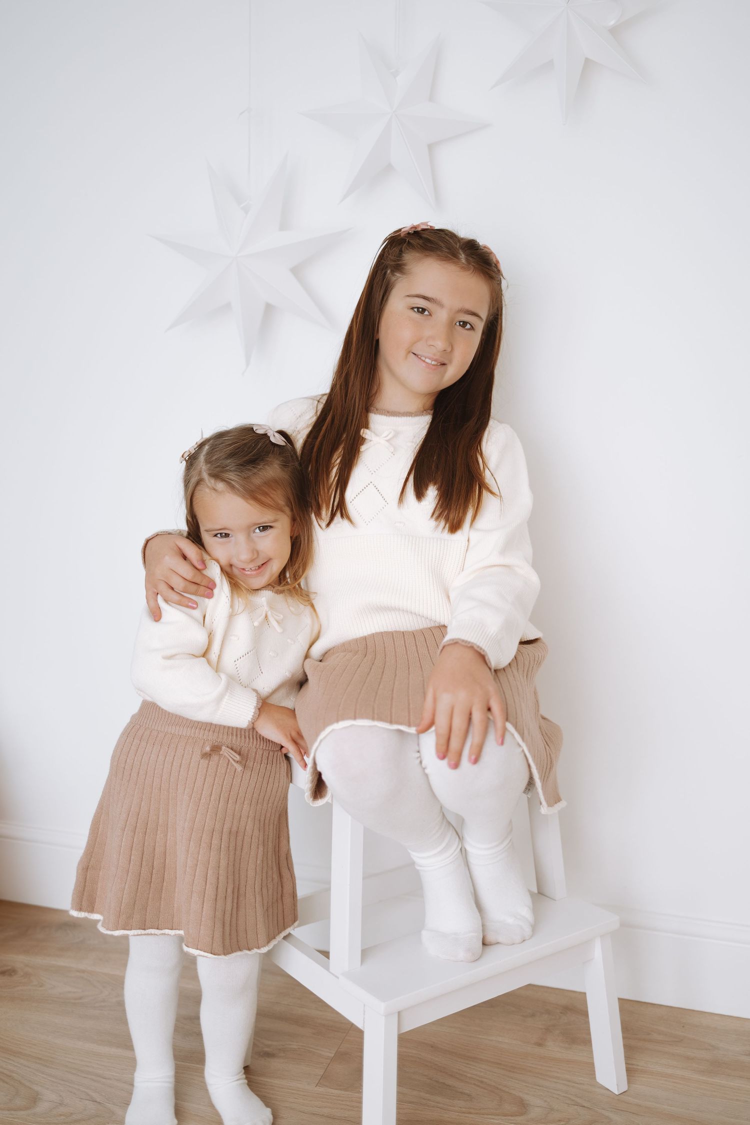 Two people in white outfits and beige pleated skirts sit together on white chairs against a light backdrop.